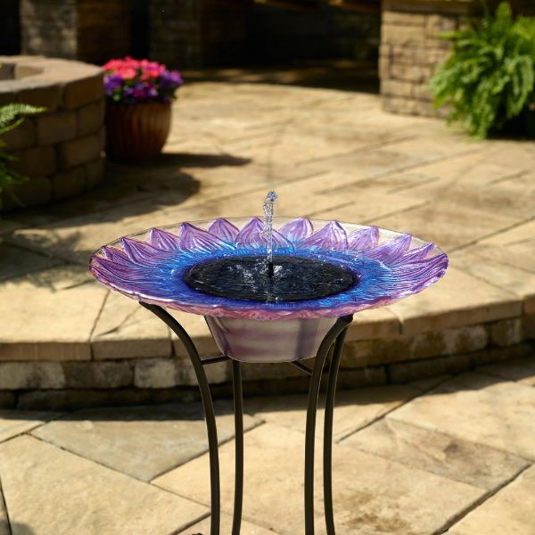 Colorful glass bird bath on a patio with flowers and a fire pit in the background