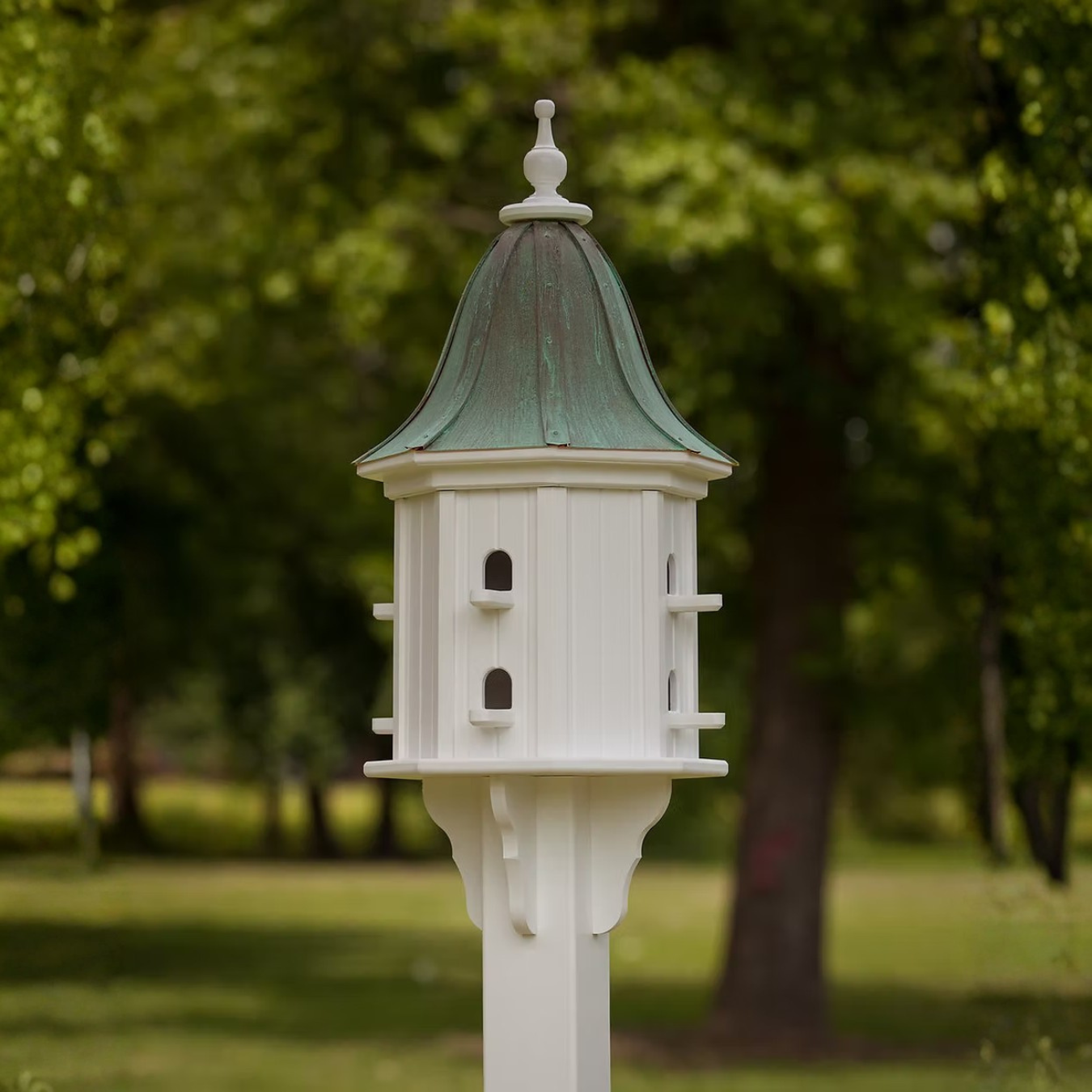 Decorative birdhouse on a stand with multiple perch entries and a bell shaped patina copper roof wuth trees in the background