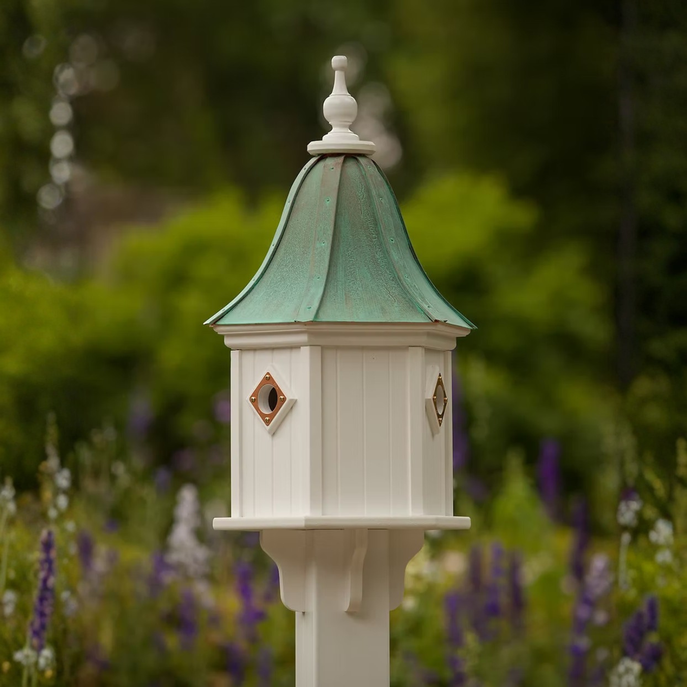 Decorative white birdhouse with a bell shaped patina copper roof in a garden setting