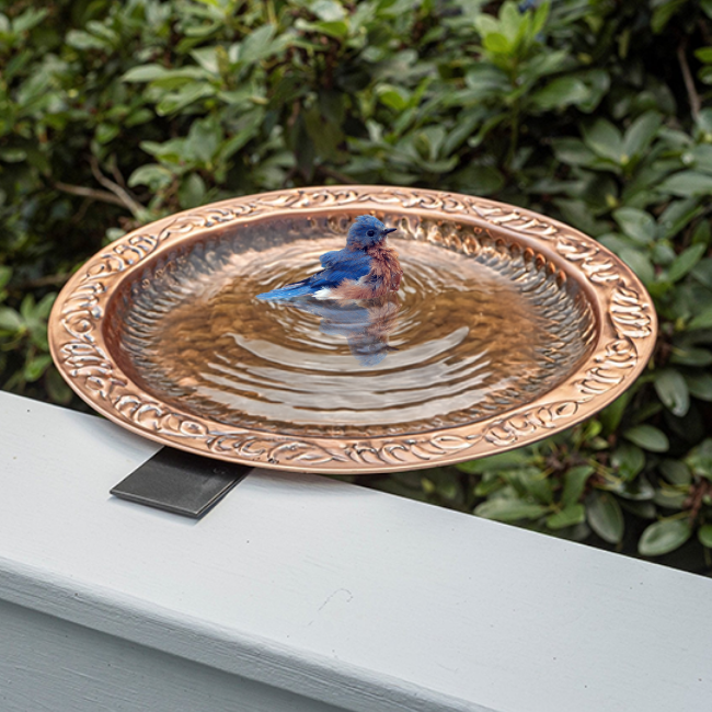 A vibrant bluebird bathing in a beautifully embossed copper bowl, mounted on a deck railing with greenery in the background.