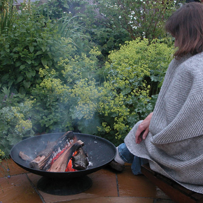 A person enjoying a fire in the Rustic Glow Firepit Bowl, sitting comfortably next to the crackling flames surrounded by greenery.