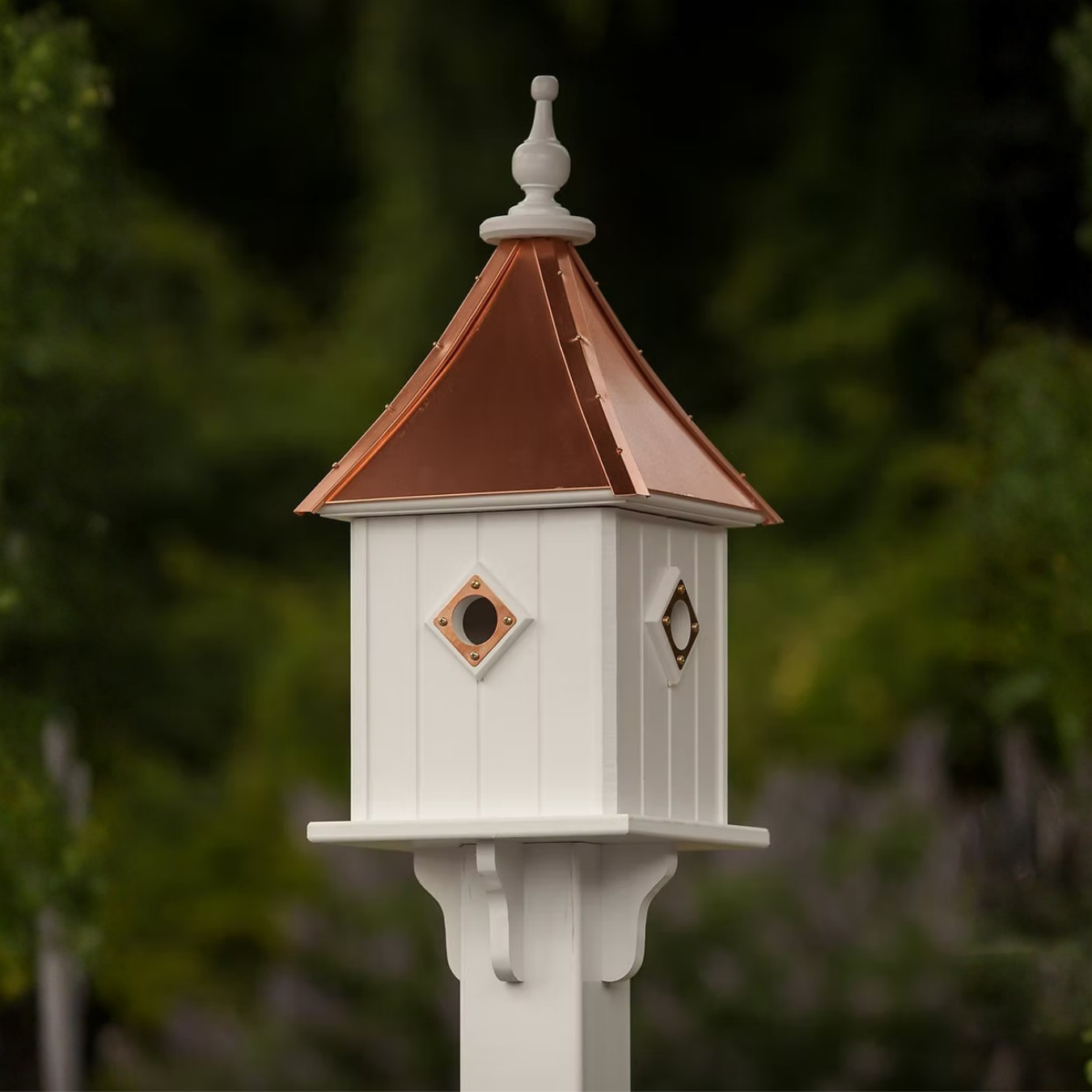 Square white birdhouse with multiple portal entries and a copper roof against a blurred green background