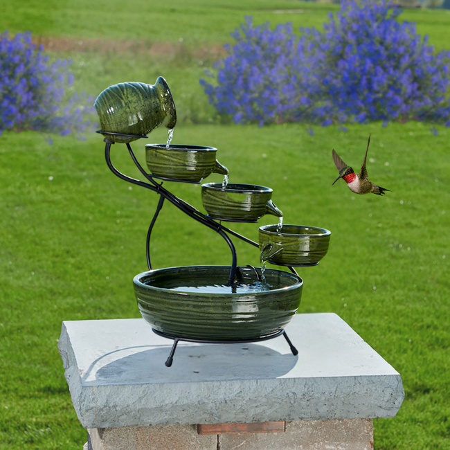 A green cascading solar fountain with 4 tiers and a hummingbird flying near it in a grassy area.