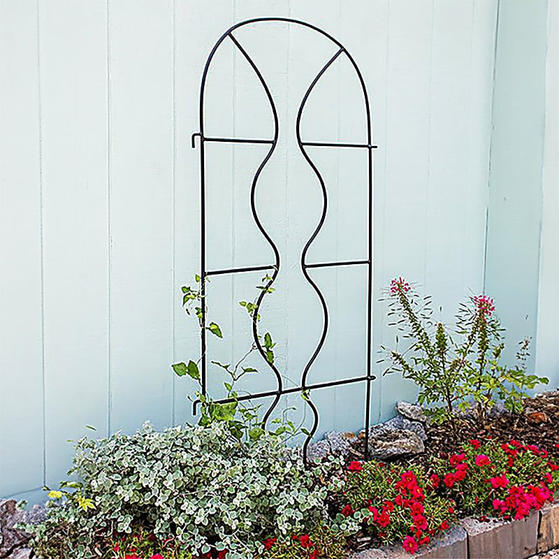 Black wrought iron arch garden trellis standing against a pale blue wall with red flowers at its base