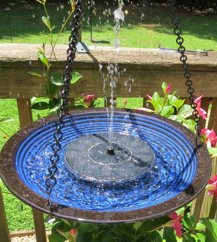 A solar-powered bird bath with a blue glazed ceramic edge and a black center, hanging from a chain, with water flowing up from the bottom creating a fountain effect.