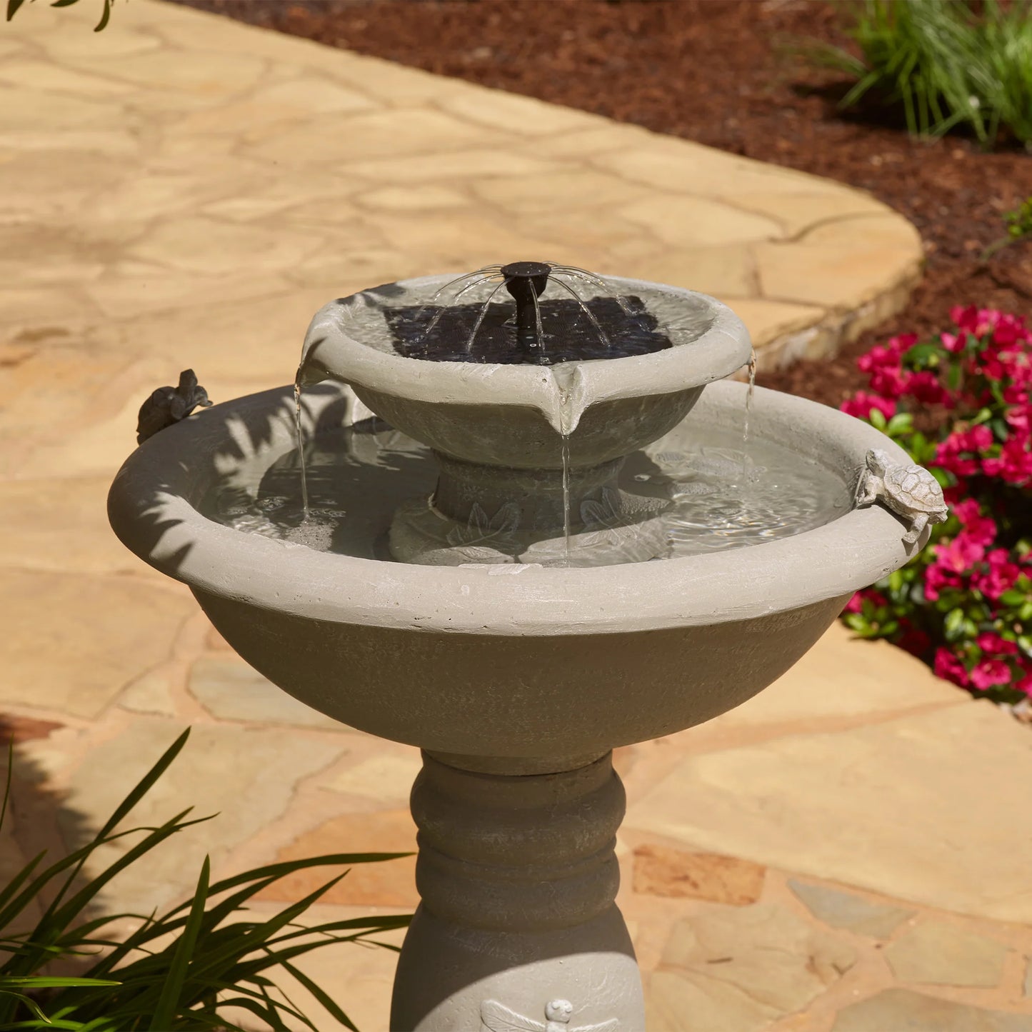 Close-up view of a two-tier outdoor fountain with a solar-powered bubbler feature, water flowing gently over the edge, surrounded by stone patio and greenery.