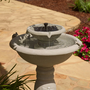 Close-up view of a two-tier outdoor fountain with a solar-powered bubbler feature, water flowing gently over the edge, surrounded by stone patio and greenery.