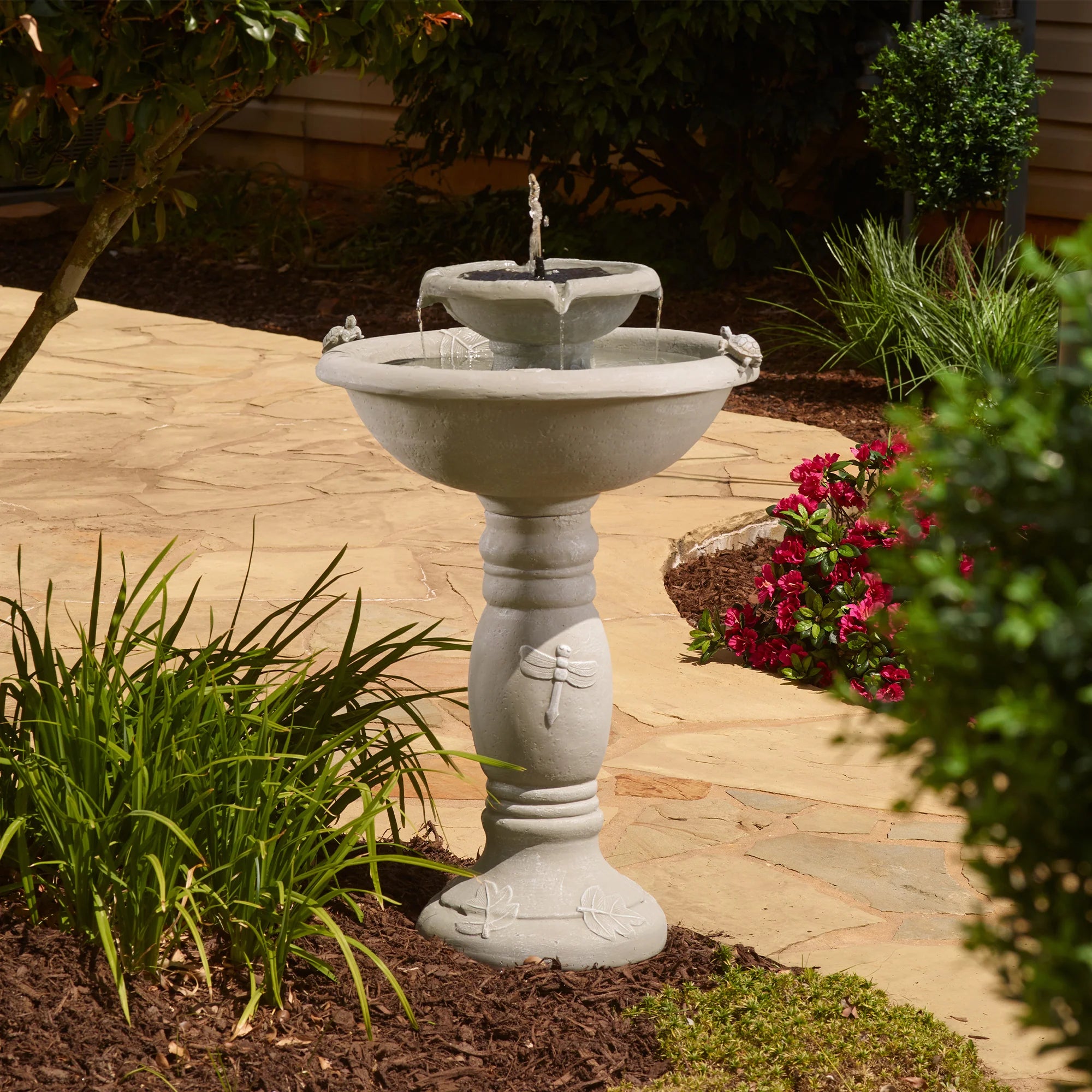 Full view of a two-tier solar garden fountain with dragonfly and leaf designs on the pedestal, surrounded by landscaped flowers and stone pathway.