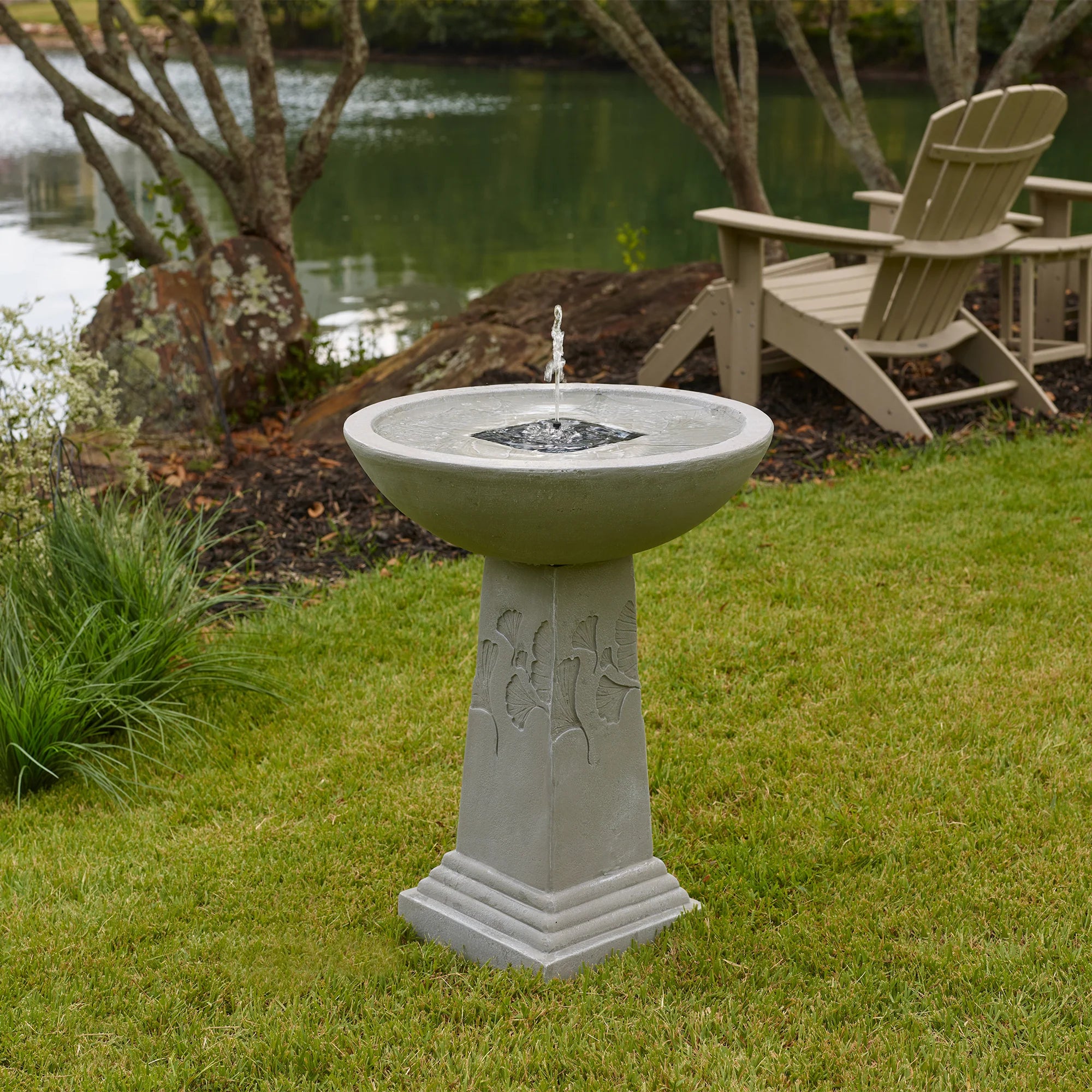 Ginko Tall Solar Bird Bath placed on a grassy lawn near a lake, with water bubbling from the fountain and Adirondack chairs in the background.