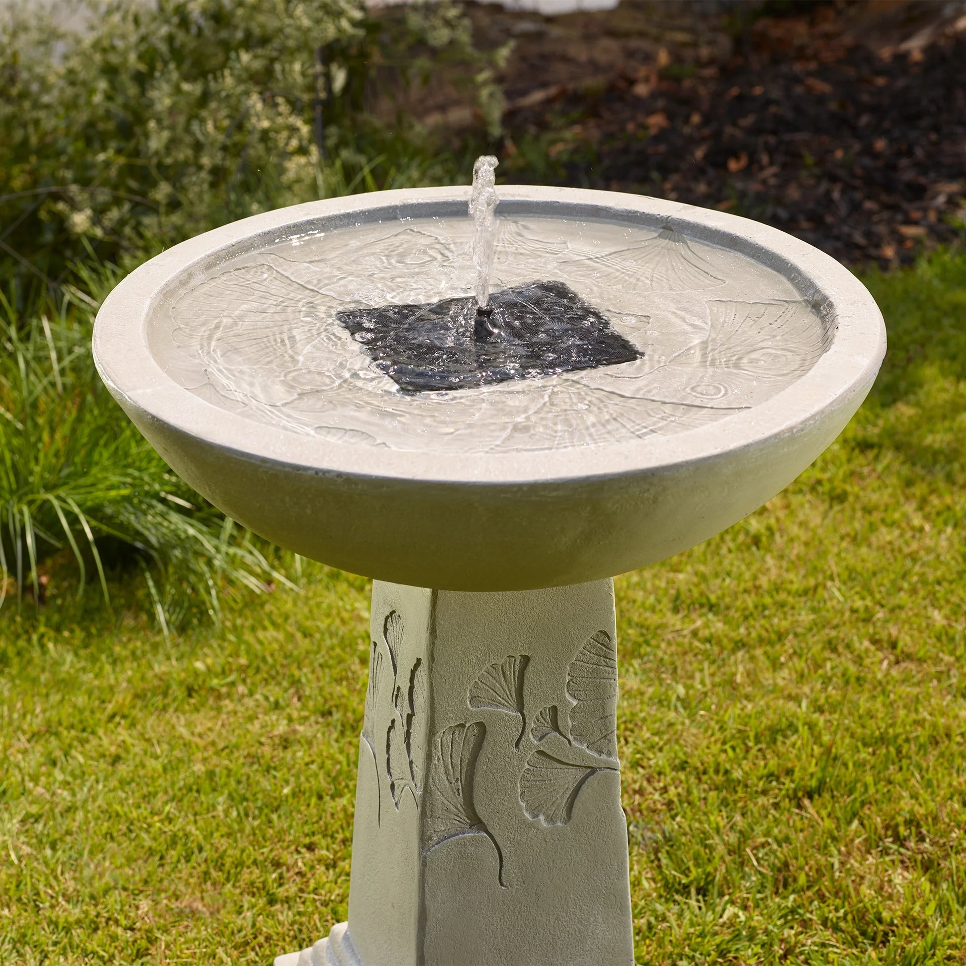 Close-up of the Ginko Tall Solar Bird Bath in sunlight, showing water bubbling from the central solar-powered fountain and intricate ginkgo leaf impressions in the bowl.
