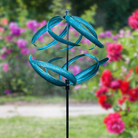 The Aqua Swirl Wind Spinner spinning gracefully in a garden with vibrant flowers, showcasing its dynamic motion and colorful design.