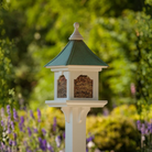 Decorative bird feeder on a stand with a Patina Copper roof and white base, set against a blurred garden background.