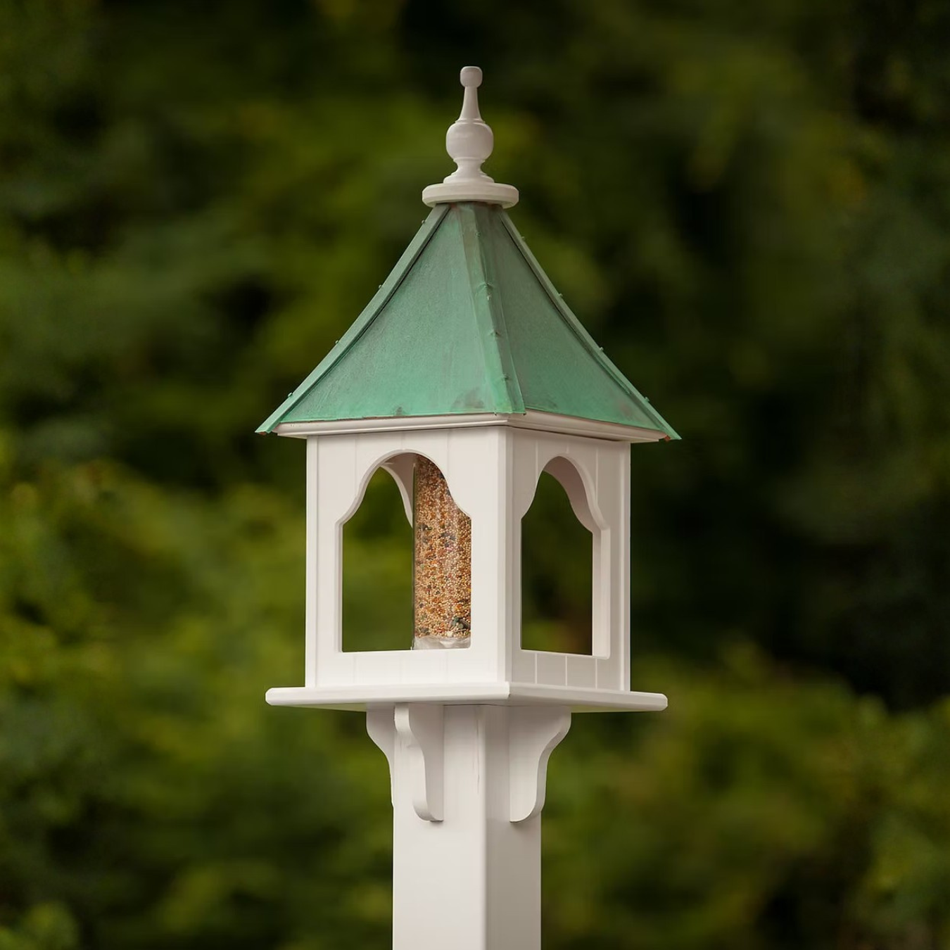 Decorative bird feeder with patina copper roof and white base against a blurred green background