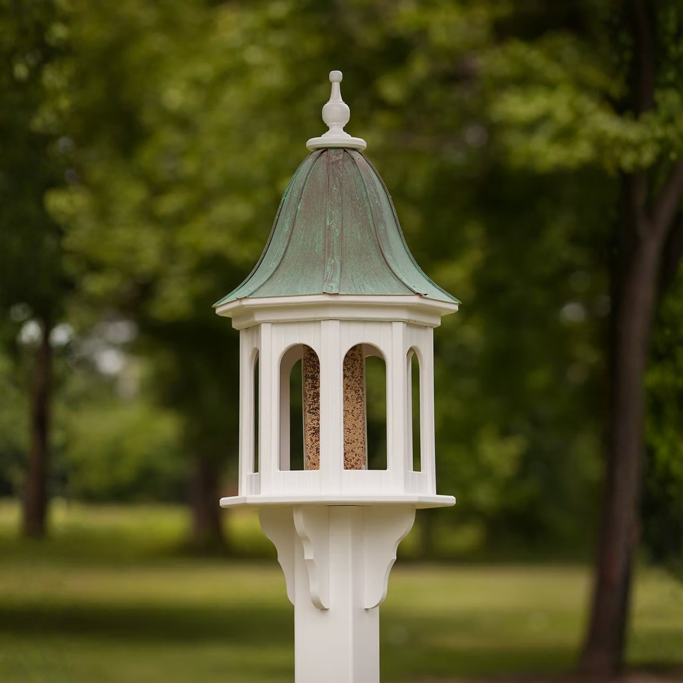 Decorative bird feeder with a patina copper roof and white base against a blurred green outdoor background