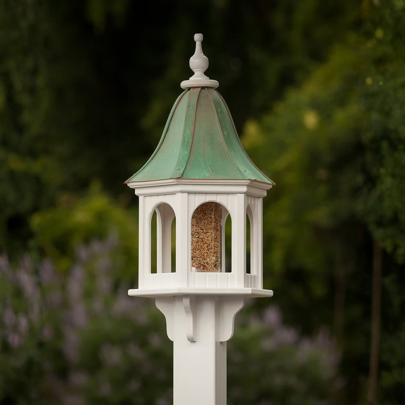 Decorative bird feeder with patina copper roof and white base against a blurred natural background