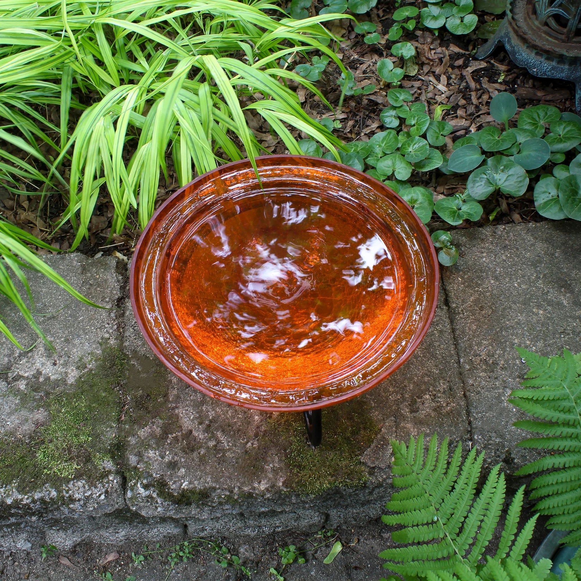 Brown ceramic bird bath on a stone patio with green plants around
