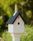 Birdhouse designed like a small church with a steeple and cross, set against a blurred green outdoor background.