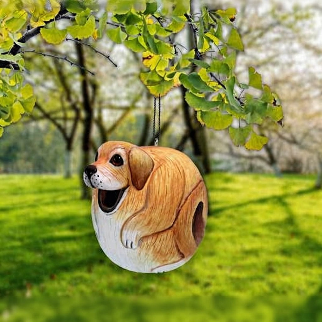 Cute birdhouse shaped like a chubby brown and white dog hangs from a tree branch with green leaves, set against a sunny, grassy park background.