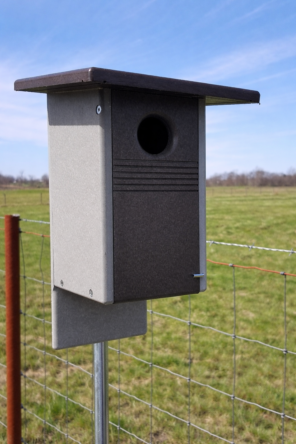Birdhouse attached to a fence post with a grassy field and blue sky in the background