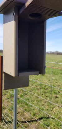 Dark-colored outdoor cat house with a wire fence and grassy area in the background