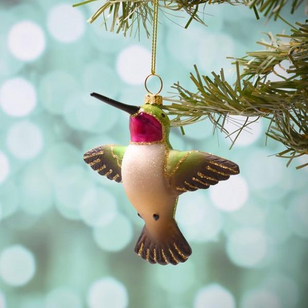 Decorative hummingbird ornament hanging on a Christmas tree branch with a blurred green background.