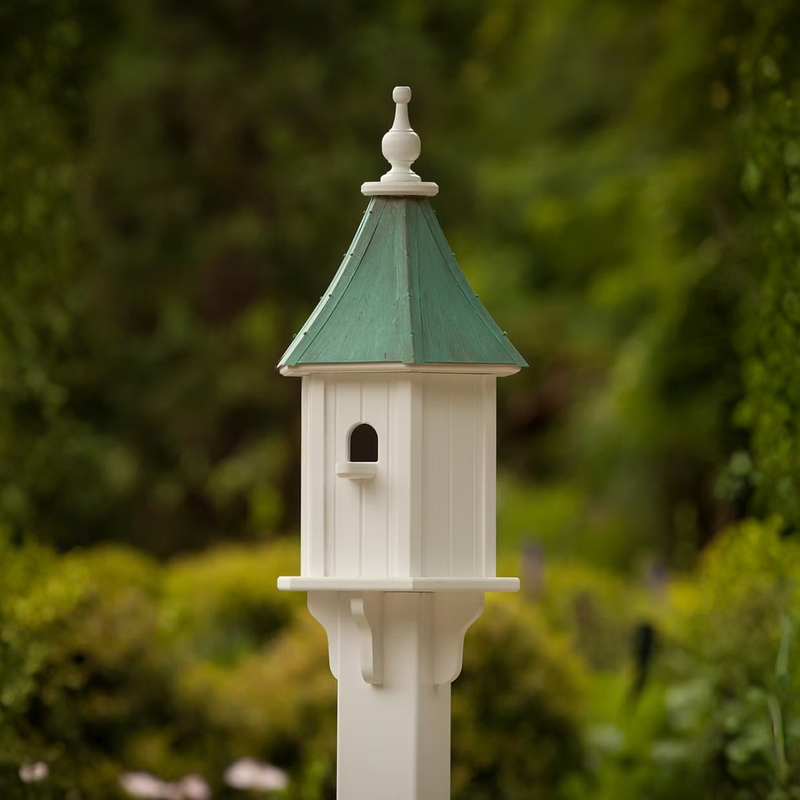 White birdhouse with perch entry and a patina copper roof against a blurred green background