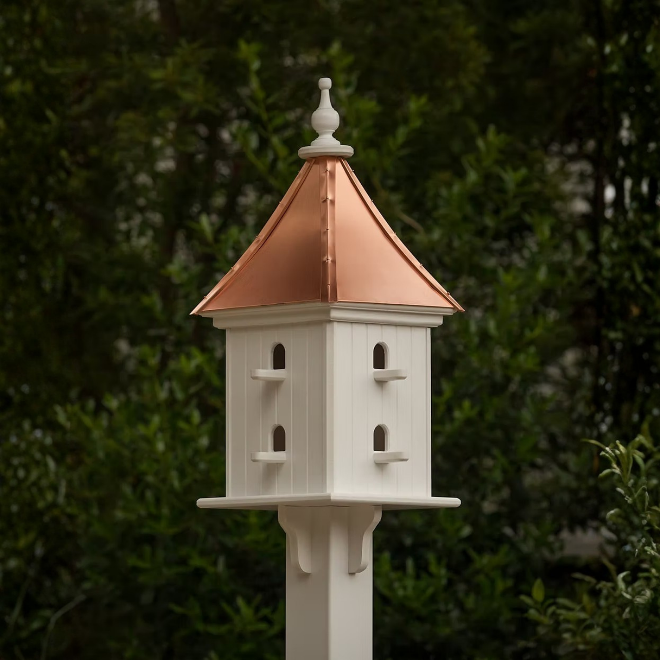 Square white birdhouse with douple perch entries on all sides with a copper roof against a green foliage background