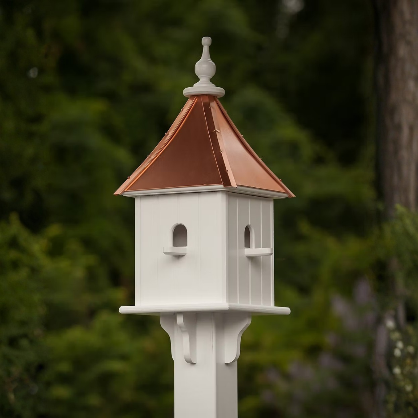 Square white birdhouse with multiple perch entries and a copper roof on a stand against a blurred green background