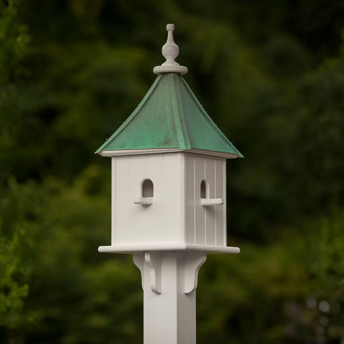 White birdhouse with multiple perch entries and a patina copper roof against a blurred green background