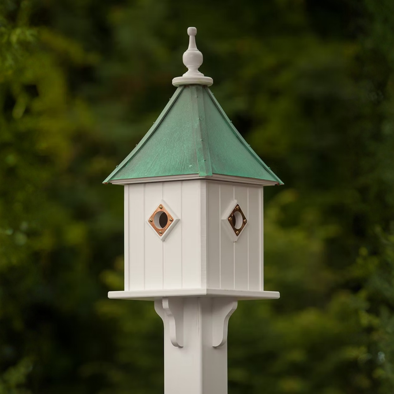 White decorative birdhouse with a sloped patina copper roof against a blurred green background