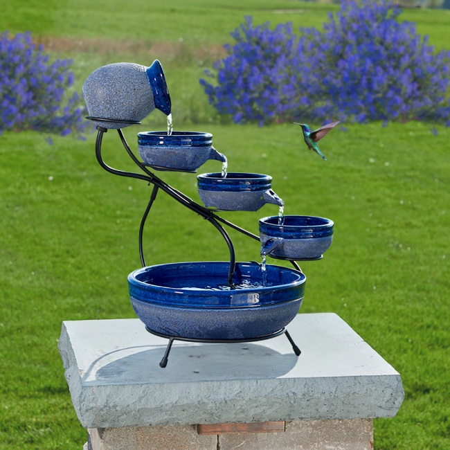 A green cascading solar fountain with 4 tiers and a hummingbird flying near it in a grassy area.