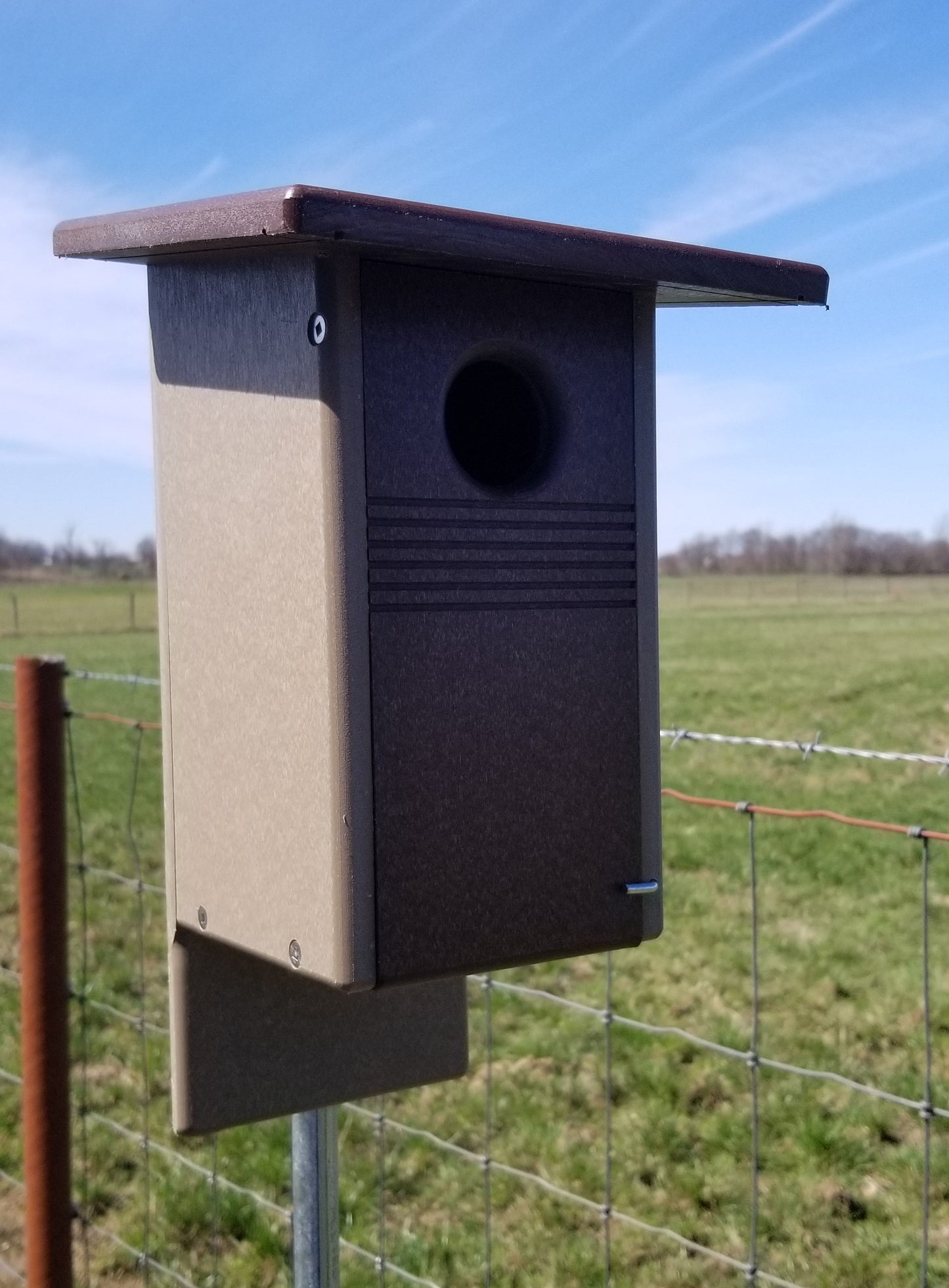 Birdhouse attached to a fence with a grassy field and blue sky in the background