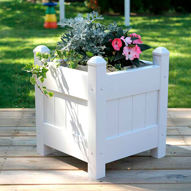 A white vinyl planter box filled with blooming pink and green plants, placed on a wooden deck for a charming garden look.