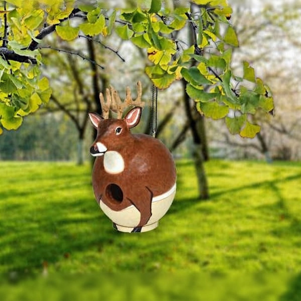 Deer-shaped birdhouse hanging from a tree branch with a green lawn and trees in the background.