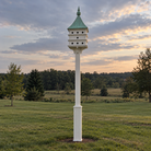 a patina copper roof purple martin house installed 10' in the air on a white vinyl post with a decorative sleeve at the base in an open backyard.